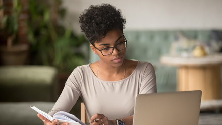 A young woman with curly hair and glasses sitting at a wooden table, working on a laptop computer. 