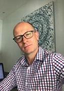 Image of a man in a check shirt and glasses, taken from a low angle. Behind him is an abstract print on the wall