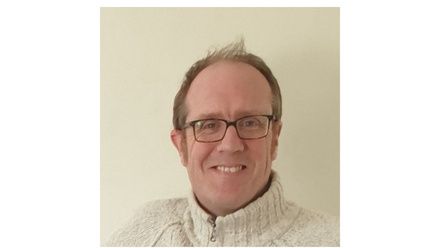 A middle-aged man with grey hair and glasses smiling at the camera. He is wearing a thick white jumper, and is against a white background 