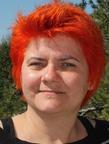 Woman with dyed red-orange hair and no make up smiling at the camera. She is outside and wearing a black top