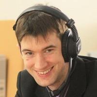 Picture of a man with brown hair smiling at the camera. He is wearing a black jacket, and also a headset 