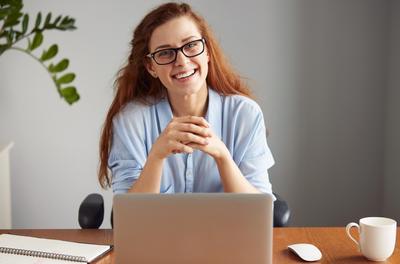 Woman with red hair and glasses, sat at a laptop, smiling 