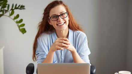 Woman with red hair and glasses, sat at a laptop, smiling 