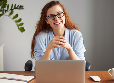 Woman with red hair and glasses, sat at a laptop, smiling 