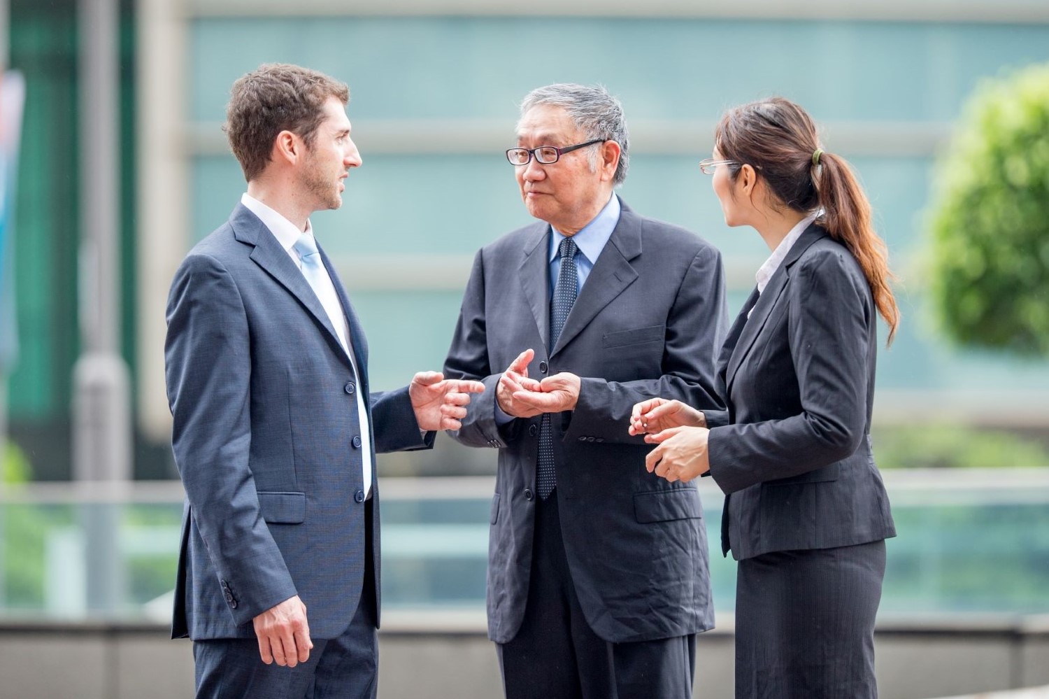 Shows three business professionals having a conversation outdoors in what appears to be a modern office or corporate environment.