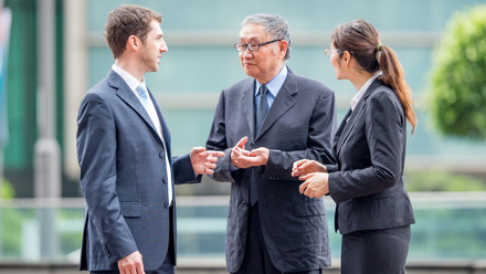 Shows three business professionals having a conversation outdoors in what appears to be a modern office or corporate environment. 