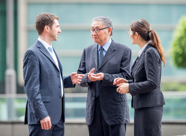 Shows three business professionals having a conversation outdoors in what appears to be a modern office or corporate environment. 