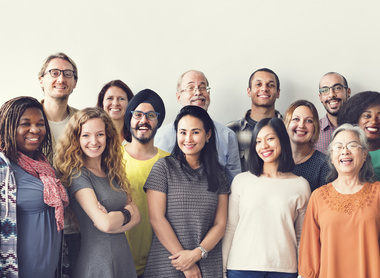 A group image of 16 people all smiling. They represent different ages, sexes and ethnicities. 
