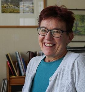 Woman with short red hair and glasses smiling. She is wearing a blue top and a white cardigan over the top