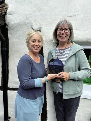 Two smiling women standing in front of a whitewashed wall, one presenting the other with a glass award.
