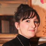 A photo of a lady with long hair styled in an updo, wearing a black high-necked top and necklace. She is smiling at the camera