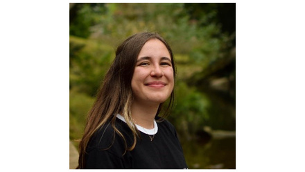 Woman with long brown hair smiling at the camera in front of some trees