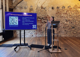 A woman presenting at the ITI French Network Workshop 2025 in a historic venue with exposed stone walls and wooden beams