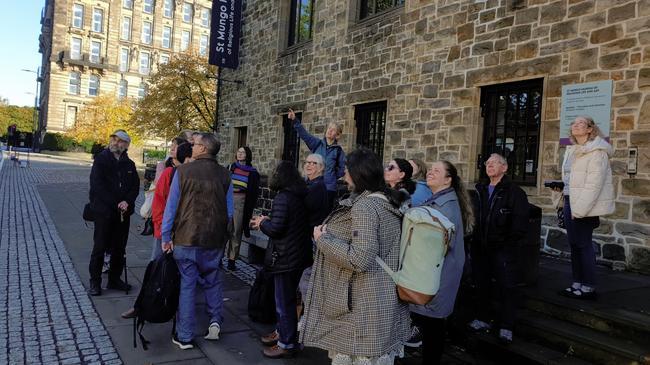 A group of people are starting on a street in Glasgow as Sara points to an architectural detail on a building