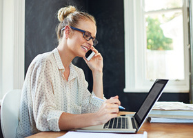 Woman sat at a laptop, on the phone, with a pen in her right hand. 
