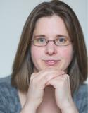 Image of a woman with shoulder length brown hair and glasses, resting her chin on her hands and smiling at the camera