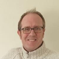 A middle-aged man with grey hair and glasses smiling at the camera. He is wearing a thick white jumper, and is against a white background 
