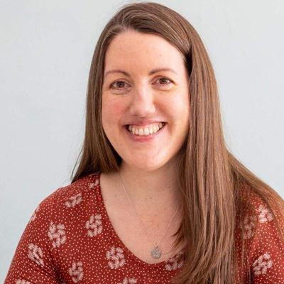 Professional headshot of a smiling woman with long brown hair wearing a red floral blouse with white polka dots and small daisy patterns.