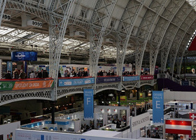 Wide-angle view of the London Book Fair inside a large exhibition hall with a glass and metal arched ceiling. The venue is bustling with attendees