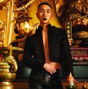 a person with short blonde hair wearing a black sequined or beaded jacket, seated in what appears to be an ornate, luxurious interior setting