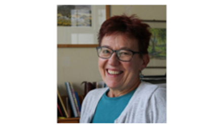 Woman with short red hair and glasses smiling. She is wearing a blue top and a white cardigan over the top 