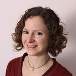 Image of a woman at 45 degrees from the camera. She has short curly hair, and is wearing a red top and a necklace