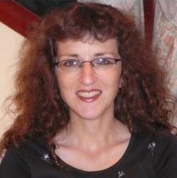 Woman with large brown curly hair and glasses, smiling at the camera. She is wearing a black top 