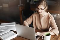 This image shows a young woman with blonde hair and glasses wearing a beige or tan colored sweater, working at a wooden table or desk. She is focused