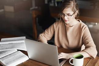 This image shows a young woman with blonde hair and glasses wearing a beige or tan colored sweater, working at a wooden table or desk. She is focused 