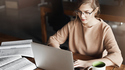 This image shows a young woman with blonde hair and glasses wearing a beige or tan colored sweater, working at a wooden table or desk. She is focused 