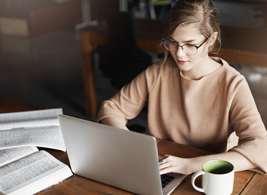 This image shows a young woman with blonde hair and glasses wearing a beige or tan colored sweater, working at a wooden table or desk. She is focused 
