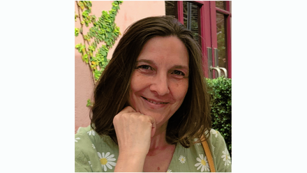 Lady with brown hair, resting her chin on her hand. She is smiling and wearing a green patterned top 